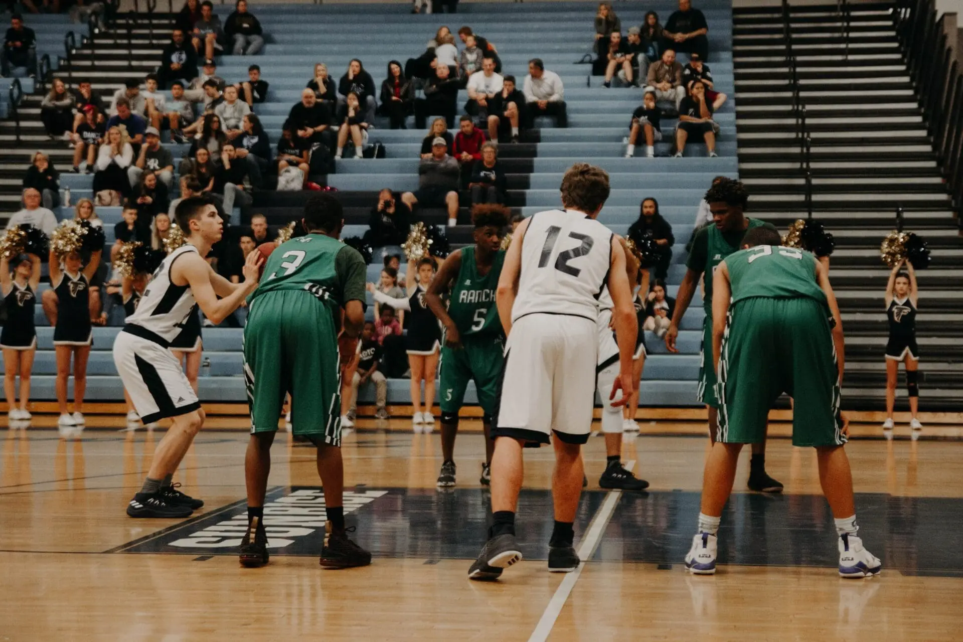 High school basketball players from two teams—one in white, one in green—prepare for a Big Time Hoops free throw on a gym court, with spectators and cheerleaders energizing the background.