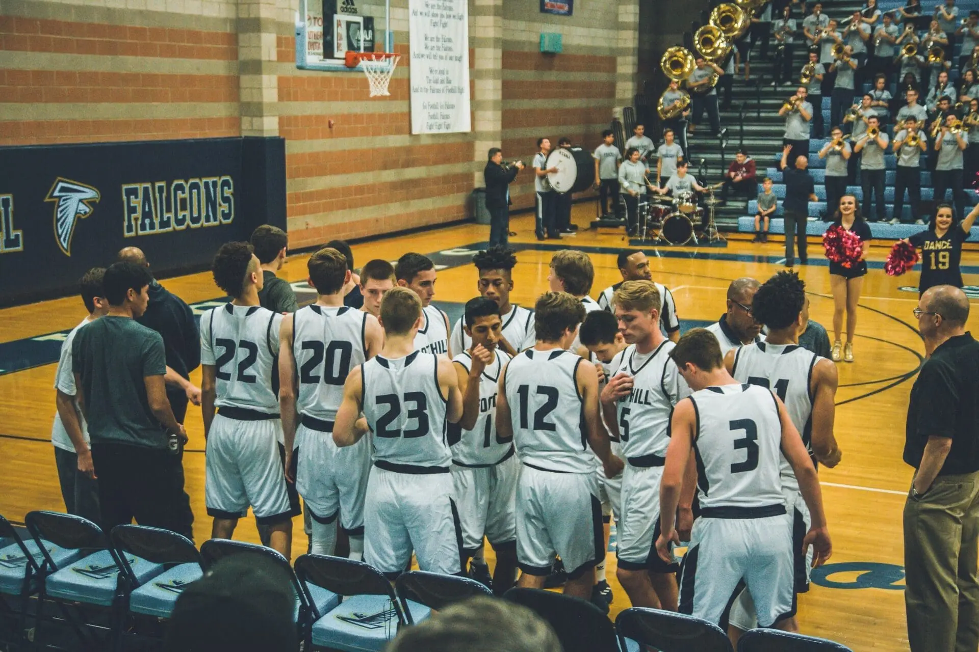 A high school basketball team in white uniforms huddles with coaches on the gym court for a Big Time Hoops game. Cheerleaders and a band perform in the background, and FALCONS is displayed on the wall.